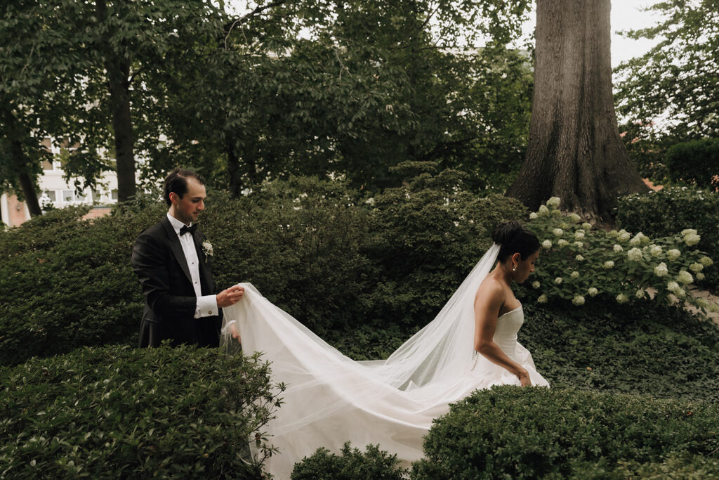 a bride walking outside with the groom behind her holding her veil and dress up